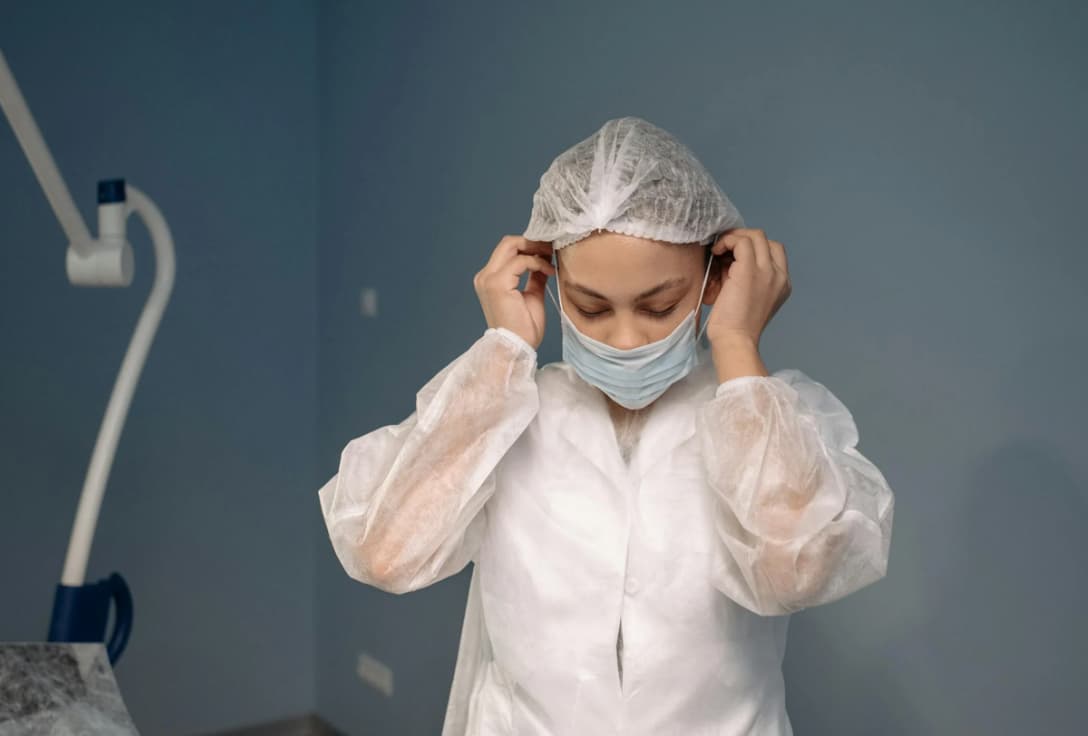 A medical professional in protective clothing adjusting a surgical cap and mask